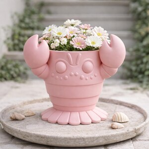 May include: A pink crab-shaped planter filled with white and pink daisies. The planter has large claw-like handles and a decorative face. The planter sits on a round, gray stone tray with seashells.