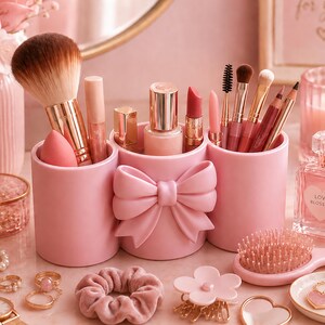 Puede incluir: Un organizador de maquillaje rosa claro con tres compartimentos cilíndricos conectados entre sí, con un detalle de lazo esculpido en la sección central. El organizador contiene varios pinceles de maquillaje, labiales y aplicadores.