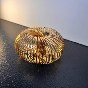 Puede incluir: Un objeto decorativo esférico dorado compuesto por anillos metálicos concéntricos. Los anillos están muy juntos, creando una estructura similar a una jaula. El objeto se encuentra sobre una superficie oscura, con un fondo degradado blanco y amarillo claro.