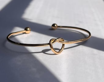 Peut inclure: Bracelet ouvert doré avec un motif de nœud en forme de cœur. Le bracelet a une finition lisse et polie et comporte de petits accents de boules à chaque extrémité. Le bracelet est présenté sur une surface blanche avec des ombres.