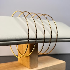May include: A set of five gold-colored bangles with a textured surface. The bangles are displayed on a white, rectangular jewelry stand with a wooden base. The bangles are thin and circular, reflecting light.
