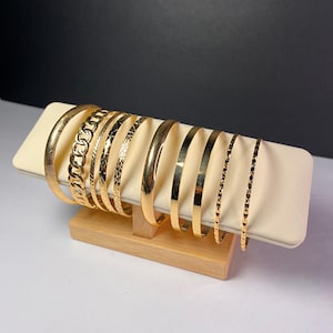 May include: A collection of gold-colored bangles displayed on a cream-colored rectangular stand. The bangles feature various designs, including chain links, textured patterns, and smooth surfaces. The stand is supported by a wooden base.