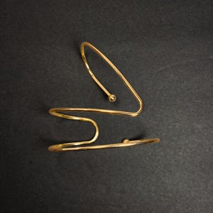 以下が含まれることがあります： 2つの金色の腕輪。片方は端に小さなボールが付いた曲線的なデザインです。もう片方は端に小さなボールが付いた曲線的なデザインです。腕輪は黒い背景に表示されています。