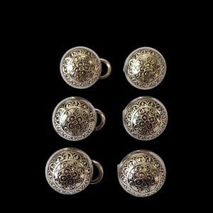Puede incluir: Seis botones plateados con un diseño floral. Cada botón tiene un pequeño lazo en el lateral. Los botones están dispuestos en dos filas de tres sobre un fondo negro.