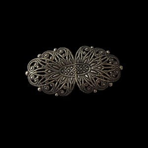Può includere: Un fermaglio in filigrana d'argento con un design delicato e intricato. Il fermaglio è di forma ovale e presenta un motivo centrale di un fiore stilizzato.