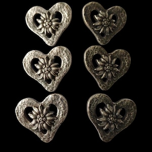 May include: Six silver-coloured heart-shaped buttons with a textured finish. Each button features a flower design in the centre. The buttons are arranged against a black background.