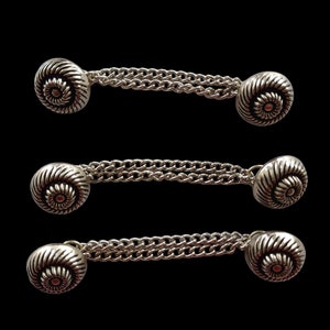 Puede incluir: Tres gemelos plateados con un conector de cadena. Cada gemelo presenta un diseño de espiral.