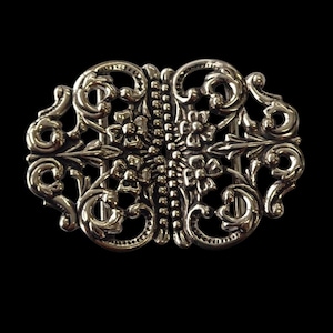 Könnte beinhalten: Verzierte silberfarbene Schnalle mit symmetrischem Design. Die Schnalle weist florale und scrollartige Details mit einer zentralen Reihe kleiner Perlen auf. Der Hintergrund ist schwarz und hebt die filigrane Metallarbeit hervor.