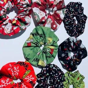May include: A collection of eight fabric hair scrunchies featuring various holiday-themed prints, including Santa Claus, candy canes, snowflakes, and green characters on red, green, and black backgrounds, arranged in a cluster on a white surface.