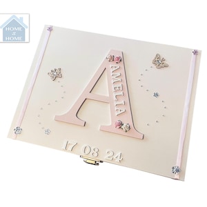 Peut inclure: Coffret souvenir rose clair avec le nom "AMELIA" et la lettre "A" en rose assorti. La bo&icirc;te est orn&eacute;e de petites fleurs, de papillons et de la date "17 08 24". Un ruban d&eacute;coratif borde les bords.