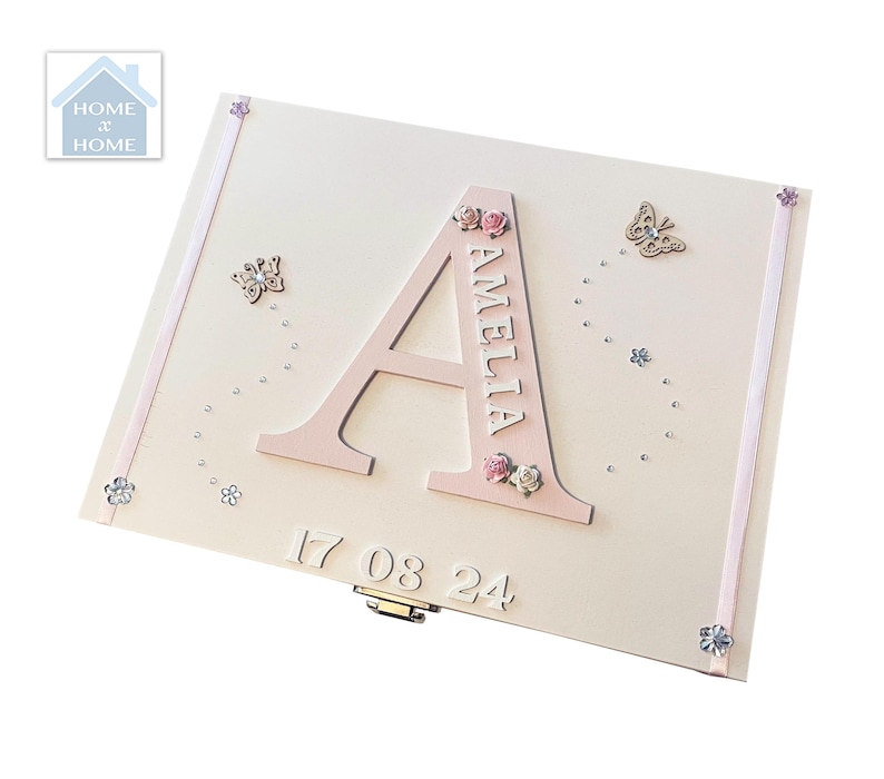 Peut inclure: Coffret souvenir rose clair avec le nom "AMELIA" et la lettre "A" en rose assorti. La bo&icirc;te est orn&eacute;e de petites fleurs, de papillons et de la date "17 08 24". Un ruban d&eacute;coratif borde les bords.
