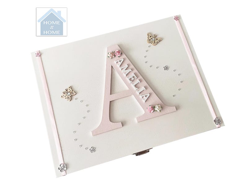 Peut inclure: Un coffret en bois blanc avec un grand "A" rose et le nom "AMELIA" en blanc. La bo&icirc;te est d&eacute;cor&eacute;e de papillons, de fleurs et de petits accents argent&eacute;s. Le logo Home x Home est visible en haut &agrave; gauche.