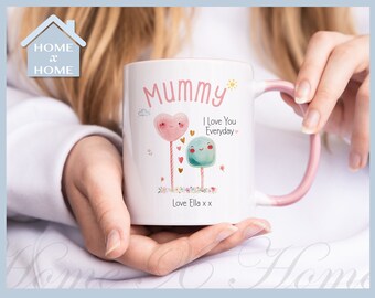 Può includere: Una tazza in ceramica bianca con un manico rosa. La tazza ha un'illustrazione colorata di due lecca-lecca con facce e il testo "Mummy I love you everyday" e "Love Ella x".