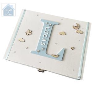 Peut inclure: Un coffret souvenir en bois blanc avec une bordure et des accents bleu clair. La bo&icirc;te pr&eacute;sente une grande lettre "L" bleu clair avec le nom "LUCAS" en blanc, ainsi que des &eacute;toiles, des nuages et des avions en bois. La bo&icirc;te est d&eacute;cor&eacute;e de petites &eacute;toiles argent&eacute;es.