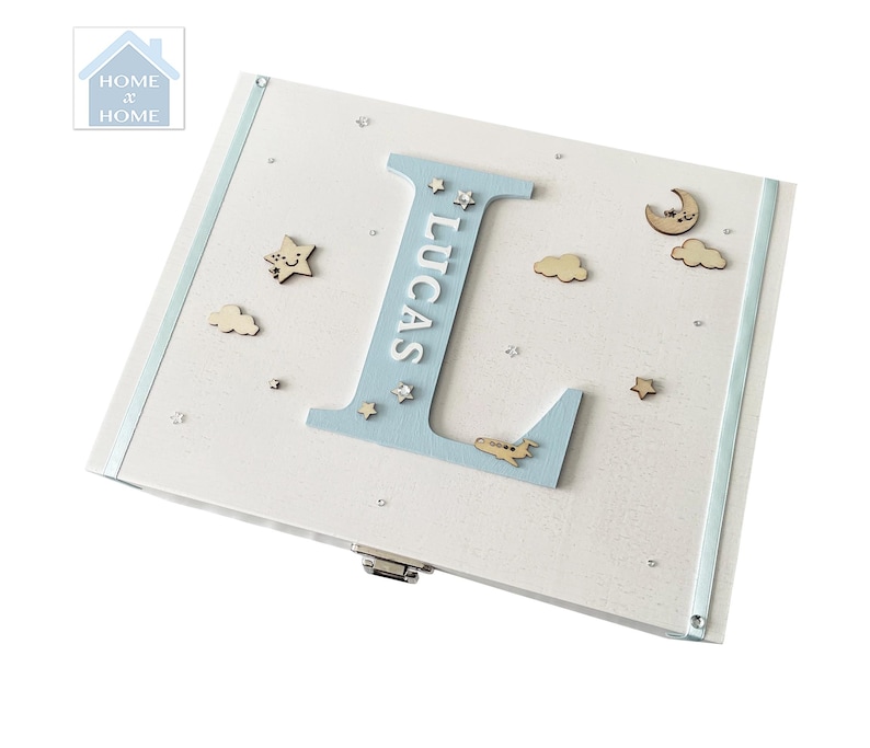 Peut inclure: Un coffret souvenir en bois blanc avec une bordure et des accents bleu clair. La bo&icirc;te pr&eacute;sente une grande lettre "L" bleu clair avec le nom "LUCAS" en blanc, ainsi que des &eacute;toiles, des nuages et des avions en bois. La bo&icirc;te est d&eacute;cor&eacute;e de petites &eacute;toiles argent&eacute;es.