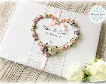 May include: White wedding guest book with a pink ribbon and a heart-shaped floral design. The heart is made of pink, white, and peach paper flowers. The text on the book reads "Chloe & James 30th March 2019".