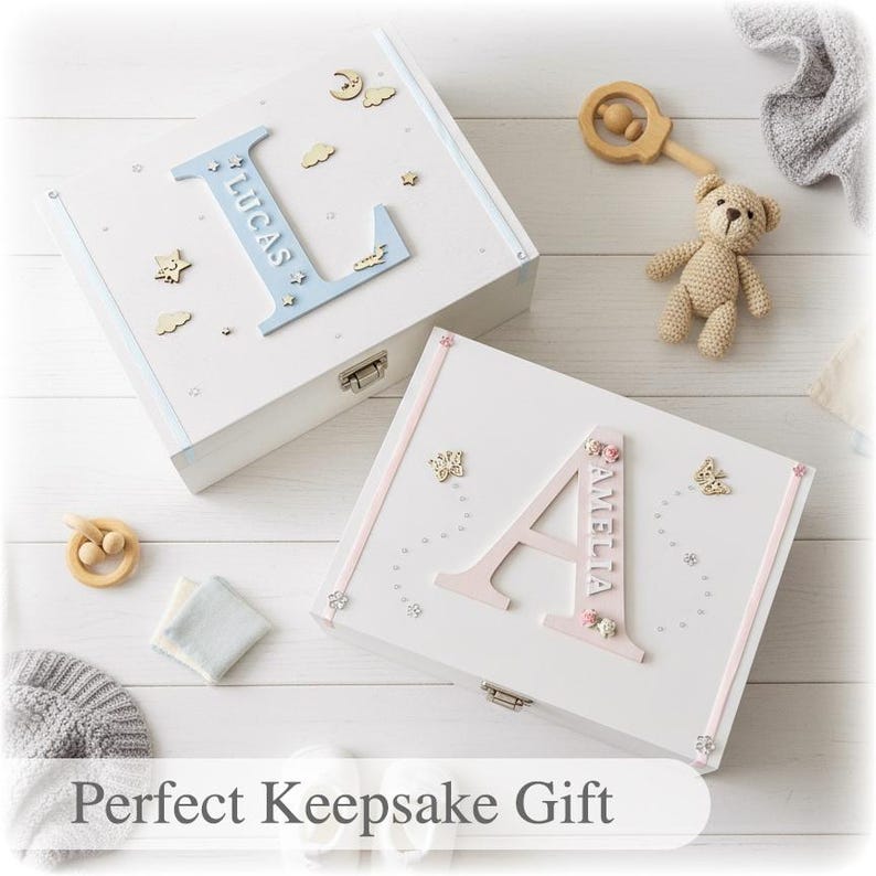 Peut inclure: Deux bo&icirc;tes &agrave; souvenirs blanches personnalis&eacute;es, l'une avec "LUCAS" en bleu et l'autre avec "AMELLIA" en rose. D&eacute;cor&eacute;es d'&eacute;toiles, de nuages et de papillons en bois. Un hochet en bois et un ours en peluche sont &eacute;galement visibles.