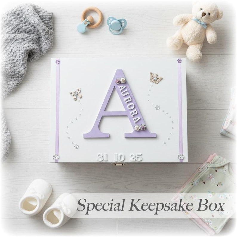 Peut inclure: Coffret souvenir blanc avec une grande lettre "A" lavande et le nom "Aurora". La bo&icirc;te est orn&eacute;e de motifs floraux et de papillons. Des accessoires pour b&eacute;b&eacute;, comme un ours en peluche et des chaussons, entourent la bo&icirc;te. Le texte "Special Keepsake Box" est visible.