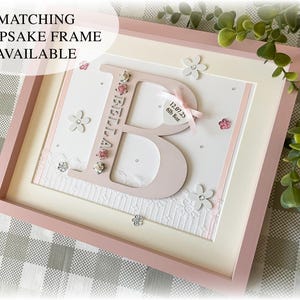 Peut inclure: Un cadre rose avec une lettre "B" d&eacute;coup&eacute;e et le nom "BELLA". Le cadre comprend des fleurs d&eacute;coratives, des strass et un ruban avec les d&eacute;tails de la naissance. Le texte "MATCHING KEEPSAKE FRAME AVAILABLE" est en haut &agrave; gauche.