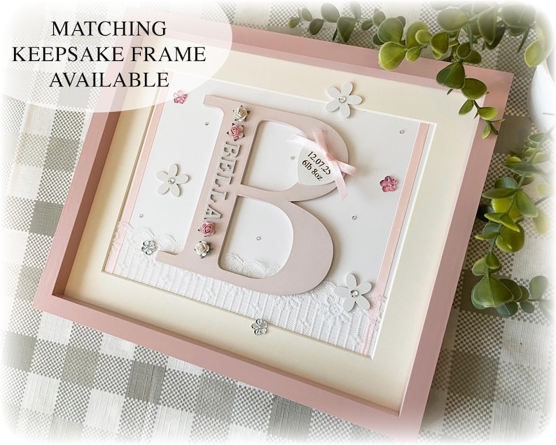 Peut inclure: Un cadre rose avec une lettre "B" d&eacute;coup&eacute;e et le nom "BELLA". Le cadre comprend des fleurs d&eacute;coratives, des strass et un ruban avec les d&eacute;tails de la naissance. Le texte "MATCHING KEEPSAKE FRAME AVAILABLE" est en haut &agrave; gauche.