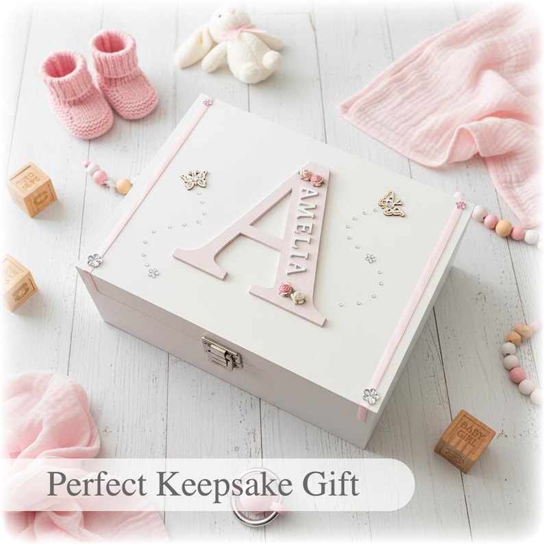 Peut inclure: Un coffret souvenir blanc avec une lettre "A" rose et le nom "Amelia". La bo&icirc;te est d&eacute;cor&eacute;e de papillons, de fleurs et de strass. Des chaussons roses pour b&eacute;b&eacute;, un lapin en peluche et d'autres articles pour b&eacute;b&eacute; entourent la bo&icirc;te.