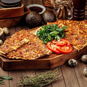 May include: A wooden platter with a variety of Turkish flatbreads topped with ground meat and spices. The platter is surrounded by fresh parsley, tomato slices, and quail eggs.