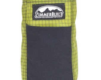 Könnte beinhalten: Eine lindgrüne Tasche mit Gittermuster, einer schwarzen Stoff-Fronttasche und einem schwarzen Kordelzugverschluss oben. Ein rechteckiger schwarzer Aufnäher mit dem weißen Text ZIMMERBUILT und einer Berggrafik ist vorne mittig angebracht.