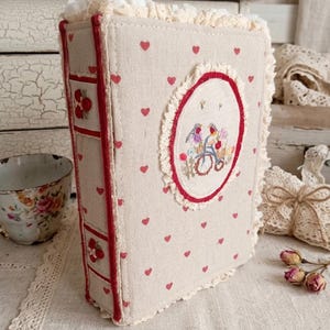 Può includere: Un diario rivestito in tessuto con sfondo beige e piccoli cuori rossi, rifinito con pizzo color crema. Il dorso presenta dettagli in tessuto rosso e ricami floreali. Un emblema ovale sul davanti mostra una bicicletta ricamata con fiori.