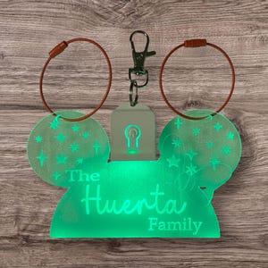 May include: A green, illuminated keychain in the shape of Mickey Mouse ears, with the text "The Huerta Family." The keychain has a silver clasp and two brown loops. The ears are decorated with stars and crosses.