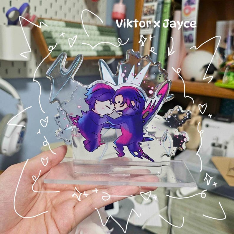 May include: A clear acrylic stand featuring a cartoon illustration of two figures embracing, with purple and pink coloring. The stand has a base and a decorative top with the text "Viktor x Jayce".