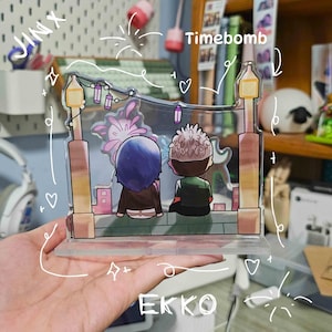 May include: A clear acrylic stand featuring an anime-style illustration of two figures seated on a ledge, with a cityscape backdrop. The stand has two lamp post designs on each side. The text "Timebomb" is visible.