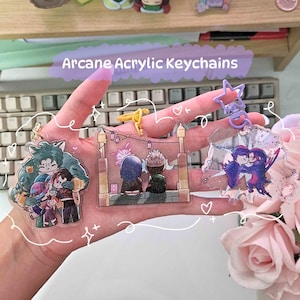 Puede incluir: Tres llaveros de acrílico Arcane con personajes del programa. El primer llavero muestra un grupo de tres personajes, uno con cabello verde y una camisa azul, otro con cabello marrón y una camisa roja, y el último con cabello azul y una camisa azul. El segundo llavero muestra dos personajes, uno con cabello azul y una camisa azul y el otro con cabello marrón y una camisa verde. El tercer llavero muestra dos personajes, uno con cabello morado y una camisa morada y el otro con cabello azul y una camisa azul. Los llaveros están todos sobre un fondo blanco.
