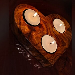 May include: A heart-shaped wooden candle holder with three lit tea lights. The holder is made of olive wood and has a natural, rustic finish.