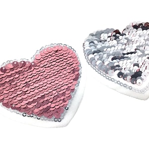 Könnte beinhalten: Zwei herzförmige Patches mit Pailletten. Ein Patch ist rosa und der andere ist silber.