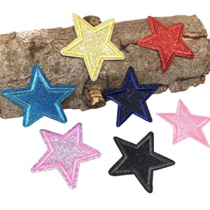 Könnte beinhalten: Sortiment von sechs Glitzer-Stern-Aufbügel-Patches in verschiedenen Farben, darunter Gold, Rot, Blau, Pink und Schwarz. Die Sterne sind auf einem Stück Holz abgebildet.