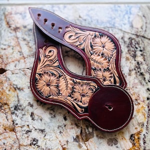 May include: A pair of dark brown leather spur straps featuring intricate hand-tooled floral and leaf patterns. The leather has a deep mahogany finish with lighter tan highlights in the carved areas, white stitching along the edges, and multiple adjustment holes.