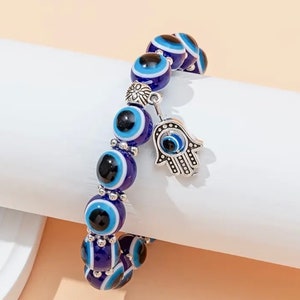 Könnte beinhalten: Blaues und weißes böses Auge Perlenarmband mit einem silbernen Hamsa-Hand-Anhänger. Das Armband besteht aus Glasperlen und hat einen silbernen Verschluss.