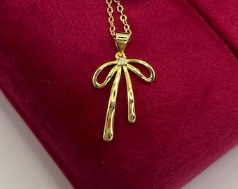 Puede incluir: Un collar dorado con un colgante en forma de lazo. El colgante tiene una pequeña gema transparente en el centro y dos líneas curvas colgantes. El collar se exhibe sobre una superficie de terciopelo rojo.