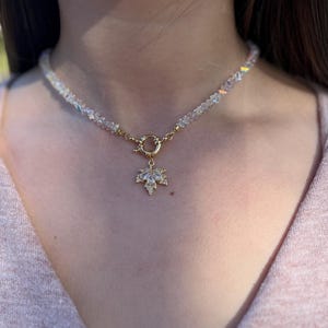 Puede incluir: Un delicado collar con cuentas de cristal iridiscentes. El collar tiene un cierre dorado y un colgante de hoja de arce adornado con cristales transparentes. La joya está diseñada para ser usada alrededor del cuello.