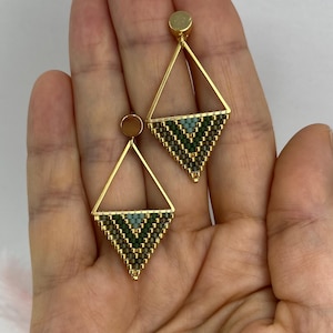 Op de afbeelding: Een paar goudkleurige driehoekige oorbellen met een geometrisch kralenpatroon. Het onderste gedeelte van elke driehoek is gevuld met kleine kralen in bosgroen, saliegroen en metallic goud, hangend aan een ronde gouden oorsteker.