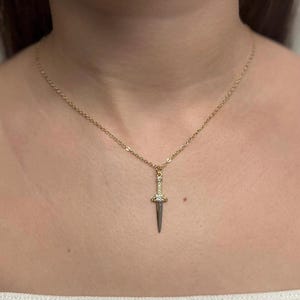 May include: A gold-colored necklace with a dagger pendant. The pendant features a small, silver-colored blade and is adorned with clear gemstones. The necklace is displayed against a skin-toned background.