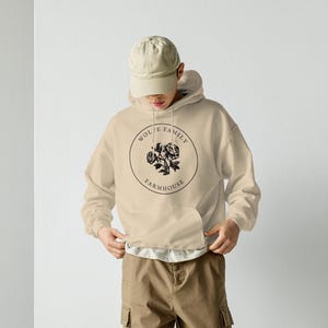 May include: A person wearing a beige hoodie with a front pocket and a black circular graphic that says "Wolfe Family Farmhouse". The person is also wearing a beige cap, brown cargo pants, and white sneakers.