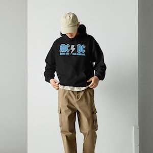 Op de afbeelding: Zwarte hoodie met de tekst "MC/DC" in lichtblauw, met een bliksemgrafiek. De hoodie heeft ook de tekst "MOTOR CITY" en "BOB CAMPBELL" in lichtblauw. De persoon draagt een beige cargobroek, een beige baseballpet en witte schoenen.