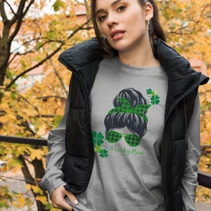 May include: A gray long-sleeved shirt with a green and black graphic of a woman with a messy bun, wearing sunglasses and a green and black plaid headband. The graphic also includes green shamrocks and the text "#LuckyMom".
