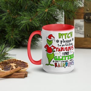 May include: A white ceramic mug with a red interior and handle. The mug features a cartoon Grinch character wearing a Santa hat and red jacket. Text on the mug includes "BITCH please," "FABULOUS," and "I PEE GLITTER."