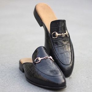 May include: A pair of black backless loafers featuring a textured crocodile-embossed pattern. Each shoe is adorned with a gold-toned metal horsebit detail across the vamp and includes a tan-colored interior lining and a low, stacked heel.