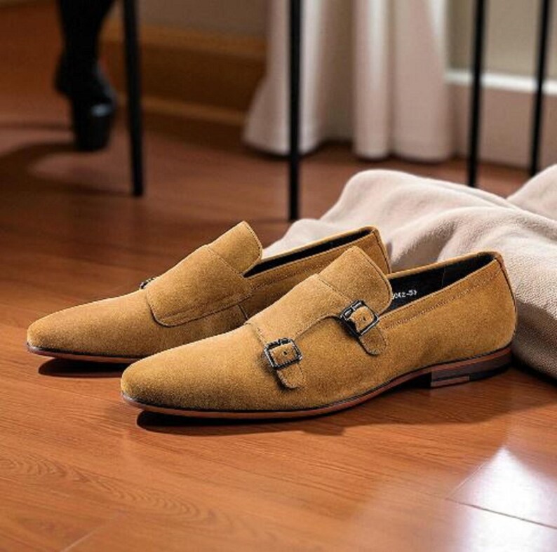 Puede incluir: Un par de zapatos de ante color marr&oacute;n claro con doble hebilla. Los zapatos tienen cierre de hebilla y est&aacute;n sobre un suelo de madera. Un estilo cl&aacute;sico, adecuado para ocasiones formales o semiformales.