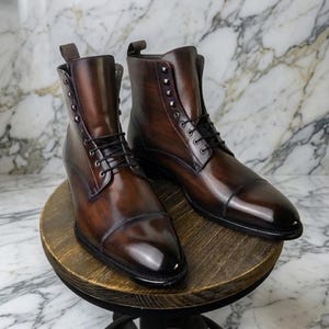 Könnte beinhalten: Ein Paar braune Lederstiefel mit hohem Schaft. Die Stiefel haben einen Schnürverschluss, eine runde Spitze und ein poliertes Finish. Die Stiefel werden auf einem Holzständer vor einem Marmorhintergrund präsentiert.