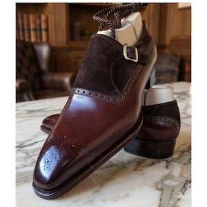 Könnte beinhalten: Ein Paar polierte, weinrote Lederschuhe mit einem dunkelbraunen Wildlederakzent am Obermaterial. Die Schuhe verfügen über einen Schnallenverschluss und dekorative Perforationen. Die Schuhe werden auf einer Marmoroberfläche präsentiert.