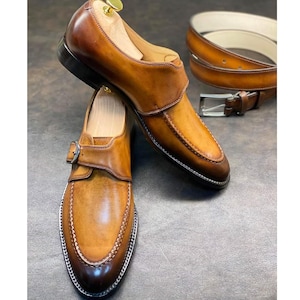 Op de afbeelding: Een paar bruinleren monk-strap schoenen met een gespsluiting en een bijpassende leren riem met een zilveren gesp. De schoenen hebben een gepolijste afwerking en decoratieve stiksels.